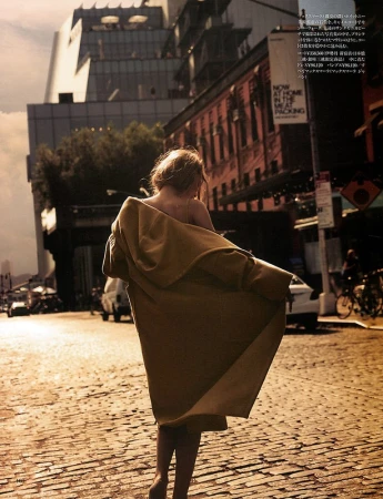 A woman with blonde hair is walking down a cobblestone street in a city, wearing a tan coat, with modern buildings in the background.