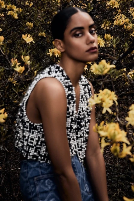 A woman in a beaded top poses amongst yellow flowers in an outdoor setting, looking directly at the viewer in a medium shot.