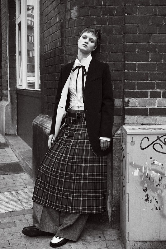 Monochrome fashion shot of a young woman posing against a brick wall, wearing a black blazer, plaid skirt, and loafers.