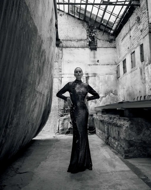 Woman in a long shimmering black dress stands in an industrial-style abandoned building, posing with her hands on her hips.