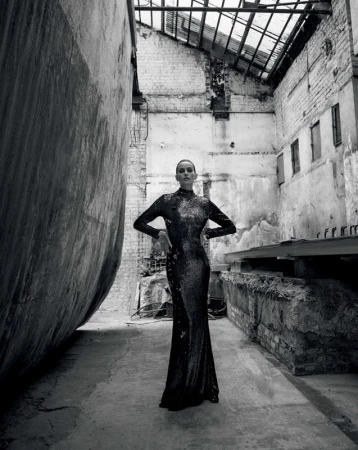 Woman in a long shimmering black dress stands in an industrial-style abandoned building, posing with her hands on her hips.