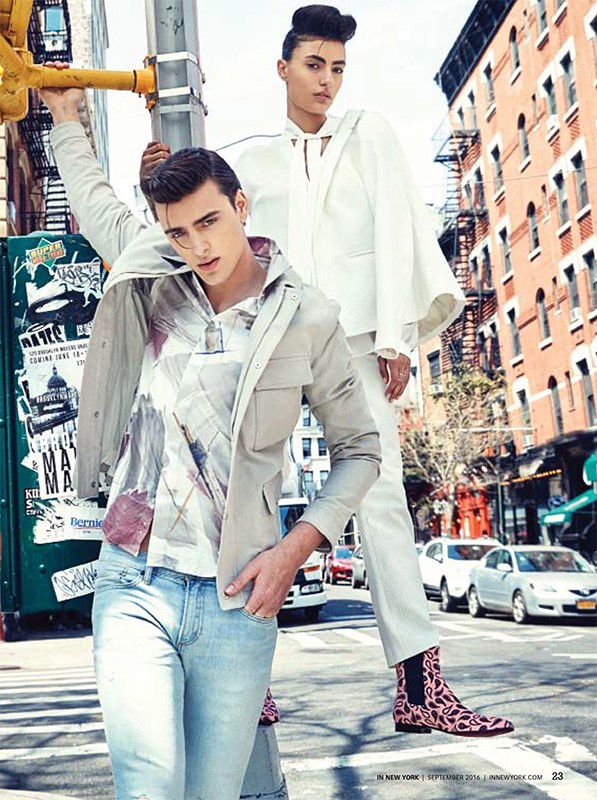 Editorial fashion shot of two models in an urban setting; a male model leans on a traffic light post while a female model stands behind him.