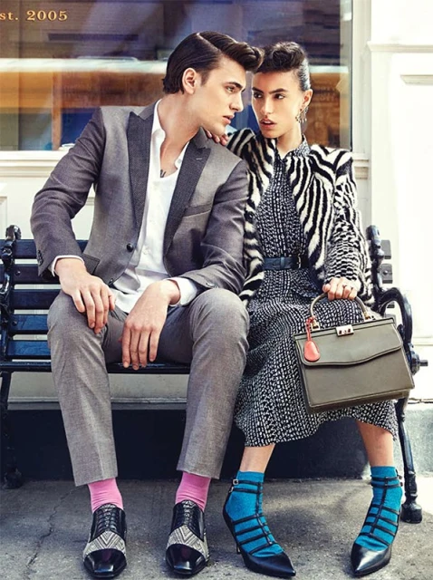 A stylish couple sits on a bench, the man in a gray suit and pink socks, the woman in a zebra-print jacket and blue hosiery.