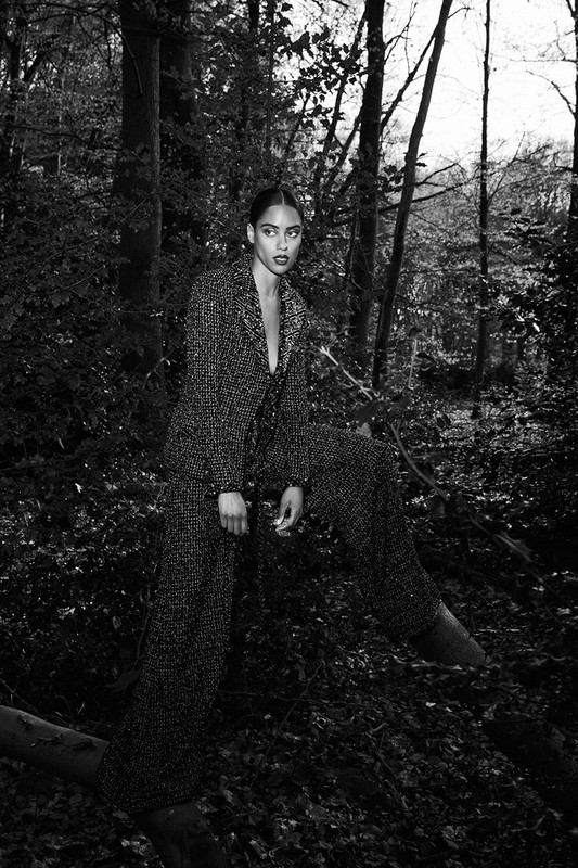 Monochrome fashion portrait of a woman standing on a fallen tree trunk in a dense forest, wearing a patterned pantsuit.