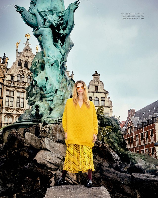 A stylish woman in a yellow sweater and patterned skirt poses in front of a vintage fountain, surrounded by classic architecture.