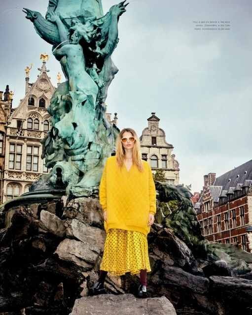 A stylish woman in a yellow sweater and patterned skirt poses in front of a vintage fountain, surrounded by classic architecture.