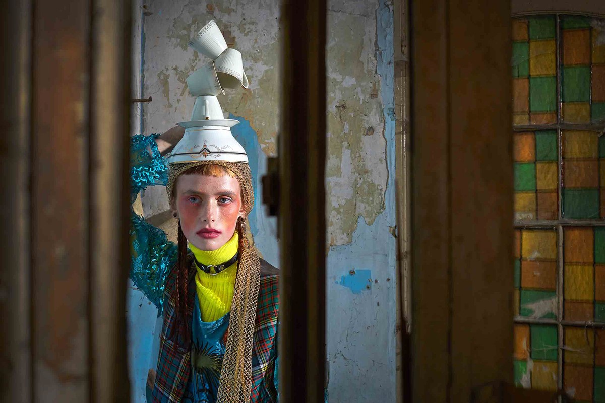 Portrait of a fair-skinned person with cups on their head, wearing a yellow turtleneck, looking through a doorway in a distressed interior.