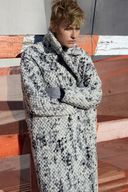 Woman poses outdoors wearing a long, patterned coat in diffused light, showcasing fashion and style in an urban environment.