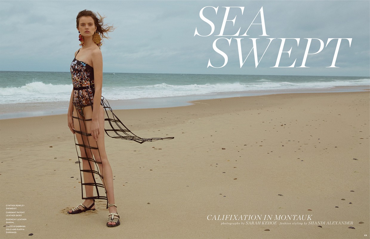 Fashion editorial image of a woman in a floral swimsuit and a skirt made of black straps, posing on a sandy beach near the ocean.