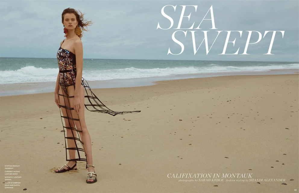 Fashion editorial image of a woman in a floral swimsuit and a skirt made of black straps, posing on a sandy beach near the ocean.