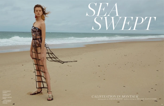 Model standing on a sandy beach wearing a patterned swimsuit and a sheer overlay skirt under an overcast sky.