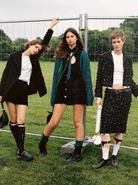 Three women in trendy outfits pose in a grassy field in front of a metal barrier, showcasing fashion and outdoor style against a natural backdrop.