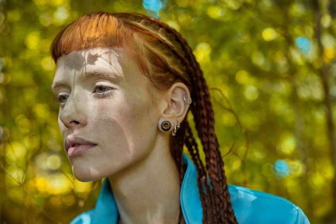 Close-up portrait of a woman with red braided hair, micro bangs, ear gauges, and a blue jacket against a leafy green background.