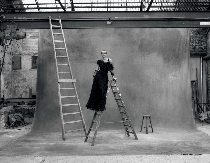 A woman in a black dress poses on a ladder in a black and white studio setting, with a stool, backdrop, and industrial details.