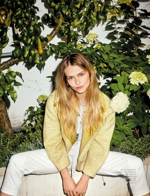 Woman with long blonde hair wearing a yellow tweed jacket and white jeans, sitting against a wall with greenery and white flowers.