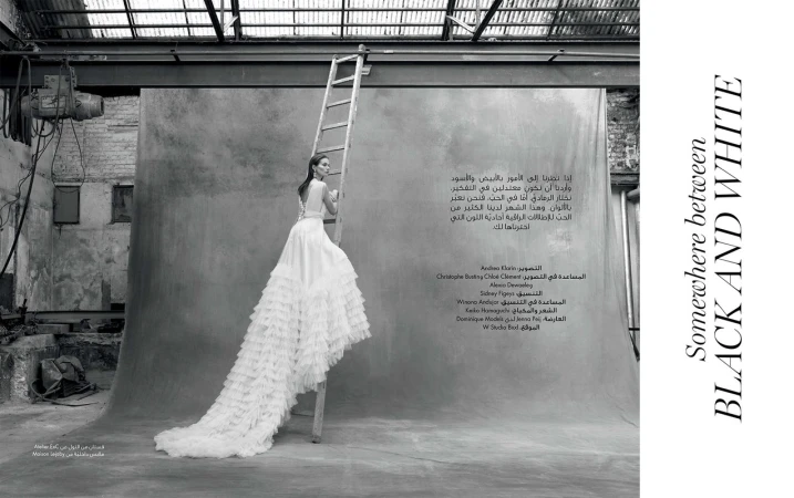 Model in white ruffled gown standing on a tall wooden ladder in an industrial studio with a textured backdrop
