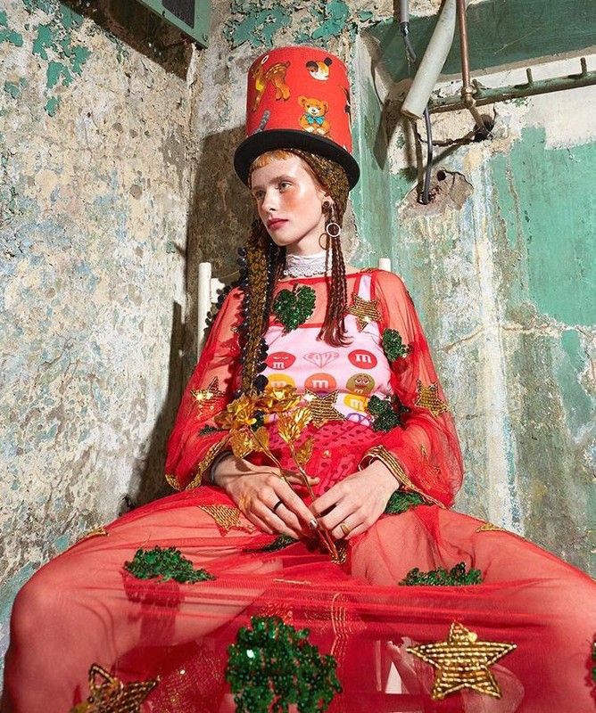 Adult woman with braids, wearing red dress and hat, sits against a distressed wall with pipes, holding gold metallic flower.