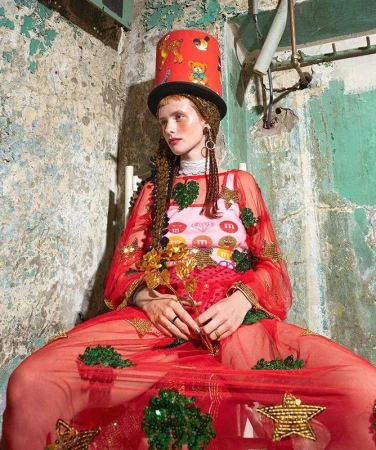 Adult woman with braids, wearing red dress and hat, sits against a distressed wall with pipes, holding gold metallic flower.
