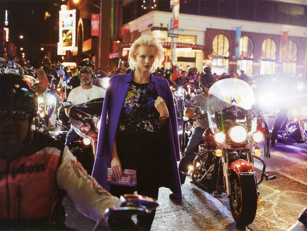 Fashion model on street with motorcycle riders at night, wearing a purple coat and holding a multicolored clutch.