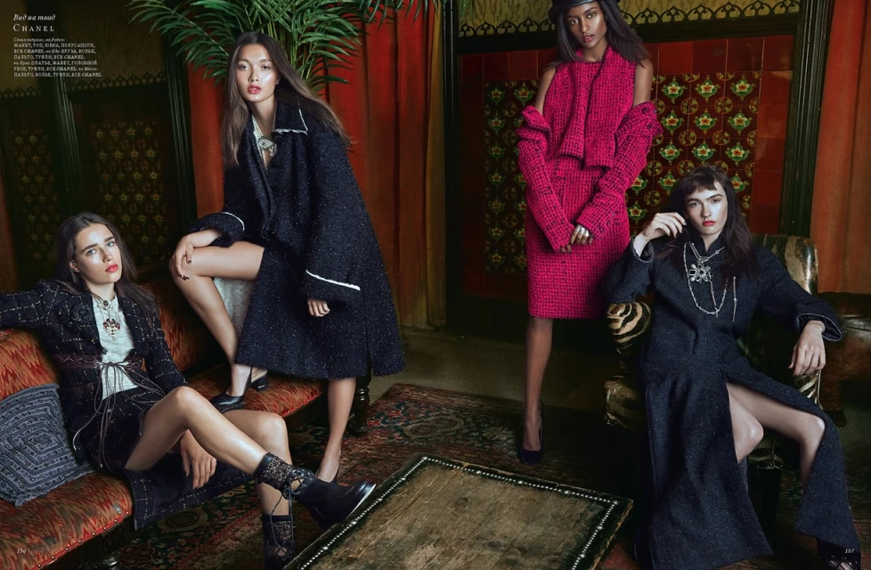 Four women in stylish, long tweed coats and dresses pose together indoors, set against a backdrop of patterned tiles and lush foliage.