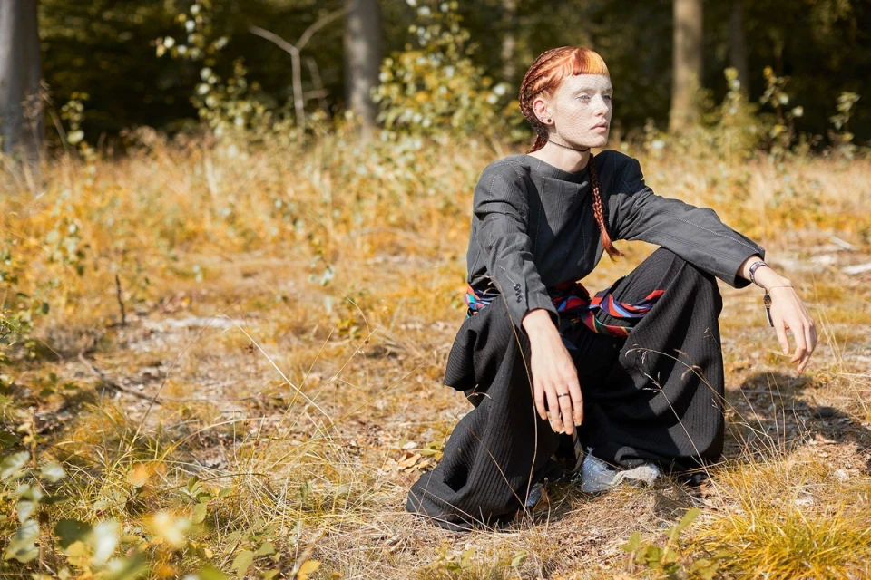 Young adult in gray top and black pants sits in a field with trees in background. Her hair is styled with red braids and short bangs.