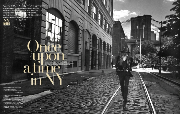 Black and white fashion editorial photo in New York City features a model wearing a dark suit on a cobblestone street near Brooklyn Bridge.