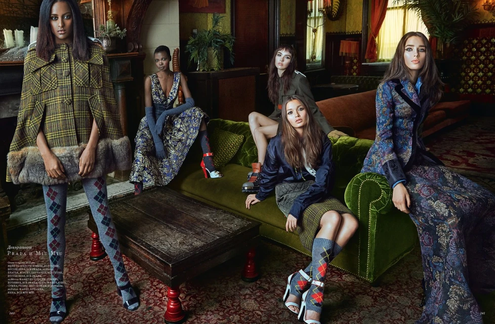 Fashion editorial shot indoors featuring five women in patterned dresses, skirts, tights, capes, blazers, and heels, posing on and around a green velvet sofa.