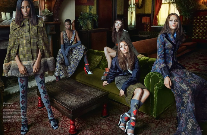 Fashion editorial shot indoors featuring five women in patterned dresses, skirts, tights, capes, blazers, and heels, posing on and around a green velvet sofa.