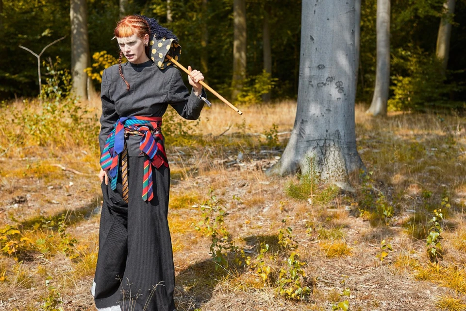 Female fashion model in the woods, wearing a grey jacket, wide black pants, and a belt made of ties, carrying an umbrella.