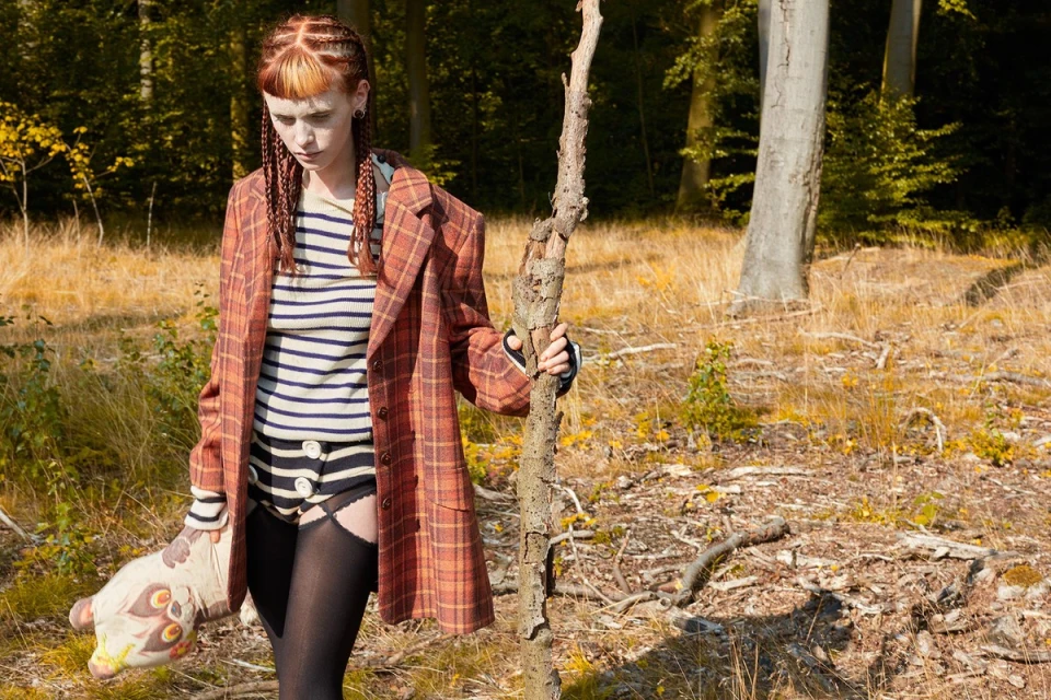 Young adult woman in woods wearing a plaid coat and holding a broken tree branch and plush toy with surreal makeup in forest setting.