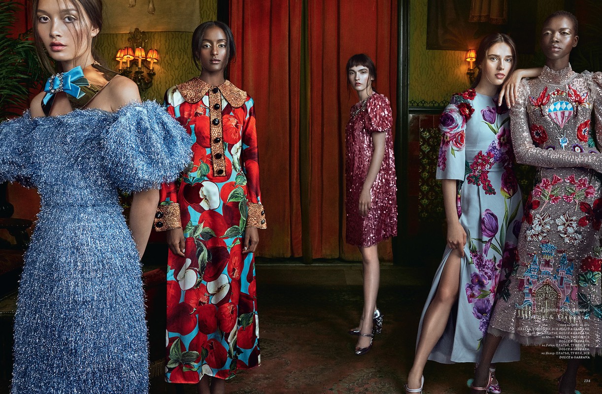 A group of five female fashion models in a dimly lit room. Each woman is wearing a uniquely designed, colorful, floor-length gown.