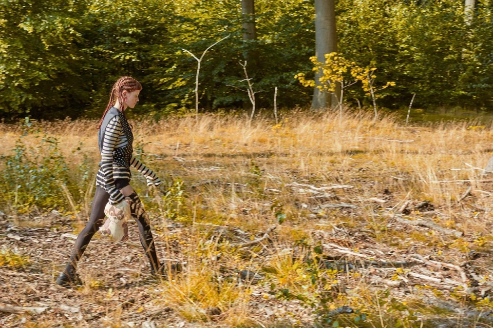 A young woman walks through a sunlit forest clearing, wearing a patterned top, leggings, and carrying a stuffed animal.