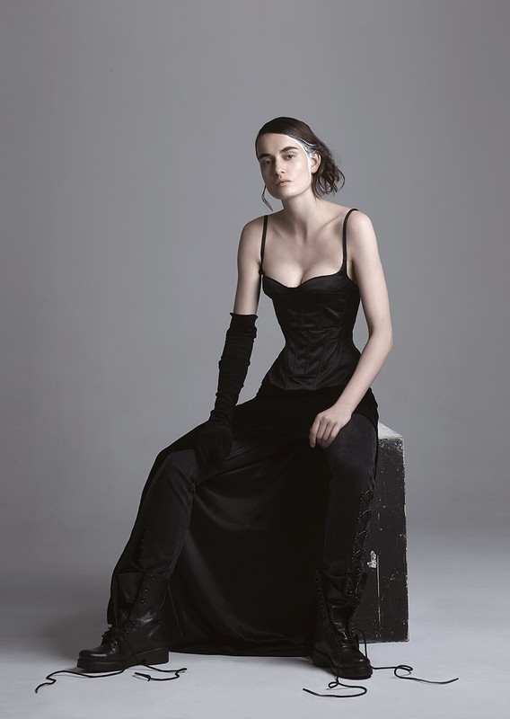 A fashion shot of a woman with dark hair in a black corset, skirt, and boots, sitting on a cube in a photo studio setting.