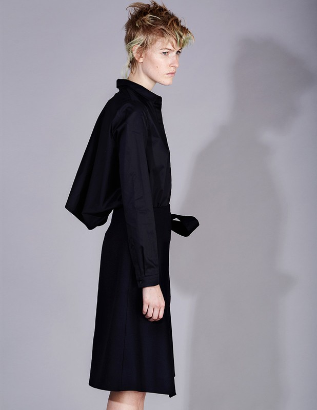 Fashionable woman in a black outfit poses in a studio with a gray background, showcasing an asymmetric dress with a tie waist.