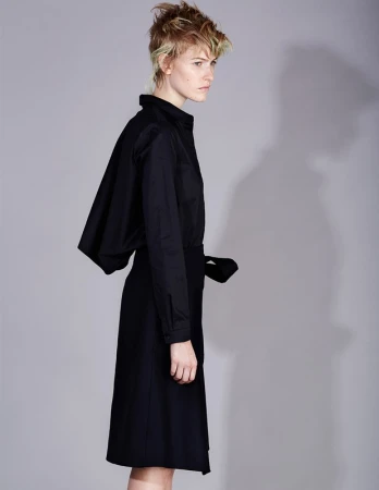 Model with short styled hair wearing a black buttoned shirt and skirt against a grey seamless studio background.