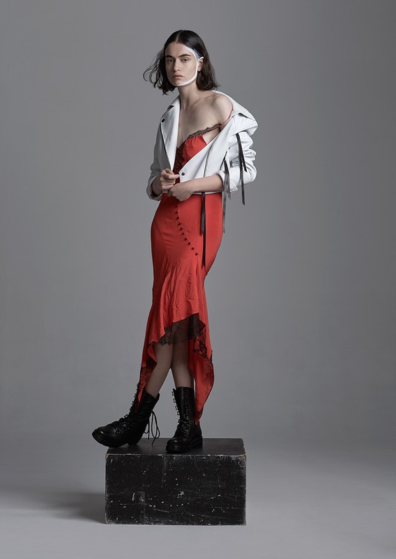 A woman in a red dress and white jacket standing on a block in a studio setting, wearing black boots and a white band around her head.
