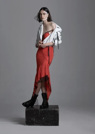 A woman in a red dress and white jacket standing on a block in a studio setting, wearing black boots and a white band around her head.