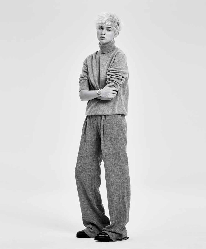 Full-length portrait of a person with blond hair, wearing a grey turtleneck sweater and wide-leg pants, standing against a white background.