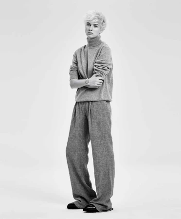 Full-length portrait of a person with blond hair, wearing a grey turtleneck sweater and wide-leg pants, standing against a white background.