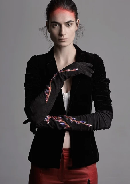 Elegant woman posing in a black velvet blazer with embellished black gloves and red trousers against a grey background in a studio setting.