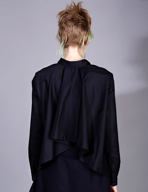 Back view of a young adult female wearing a black long-sleeved shirt and dark skirt, styled with short blonde hair with green highlights against grey.