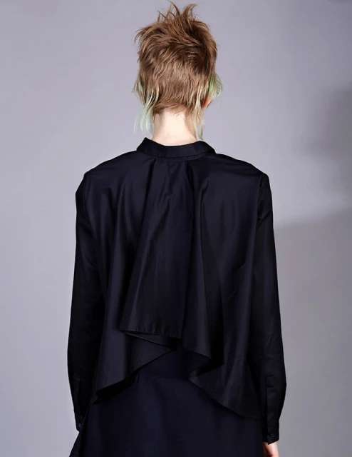Back view of a young adult female wearing a black long-sleeved shirt and dark skirt, styled with short blonde hair with green highlights against grey.