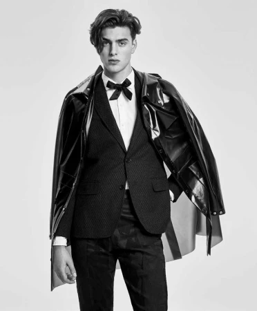 Monochrome portrait of a young man wearing a suit, bow tie, and a glossy dark raincoat draped over his shoulders.