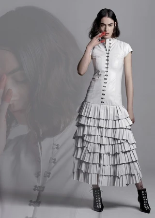 Model in white dress with tiered pleated skirt and black lace-up boots against a grey background