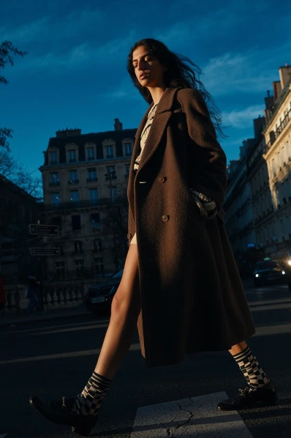 A woman in a brown coat confidently strides across a crosswalk in an urban setting, with buildings visible under a blue sky.