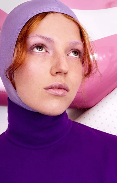 Close-up portrait of a woman with red hair wearing a purple turtleneck and hood, looking up and away from the camera in a studio.