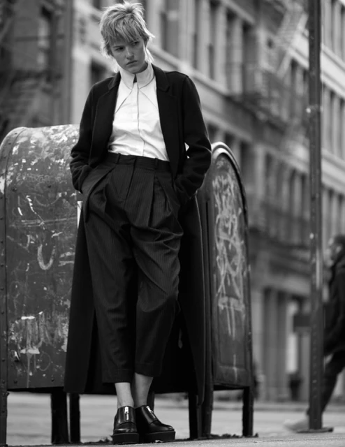 A lone woman in a dark coat stands in an urban street next to a metal box; monochrome shot.