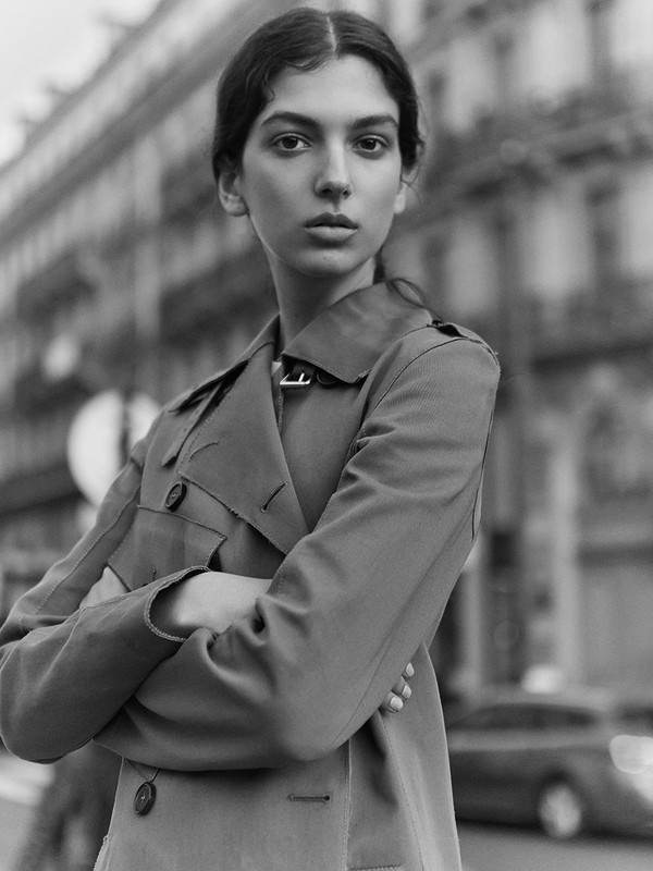 Monochrome portrait of a woman in a trench coat on a city street with buildings and a car in the background, captured in black and white.