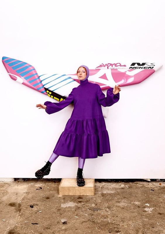 A fashion photo featuring a model in a purple dress and hat, posing on a platform in front of surfboards mounted to a white wall.
