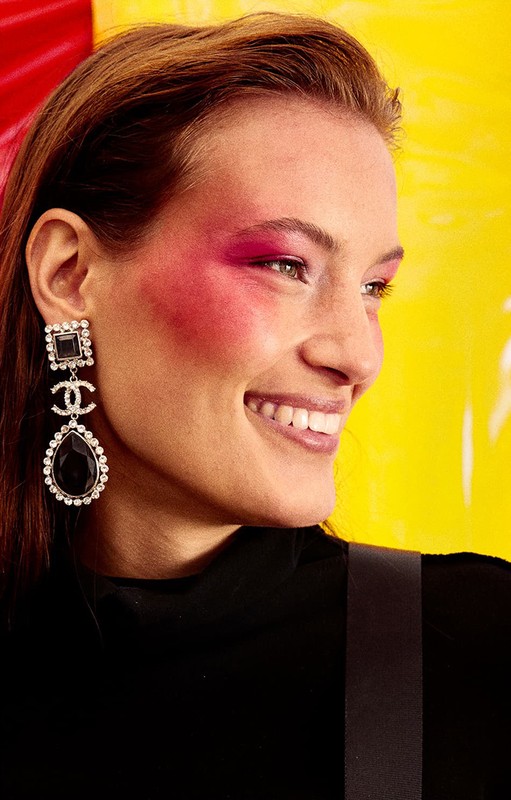Woman with pink blush and black Chanel earrings smiles against a yellow and red background in an indoor, close-up fashion shot.